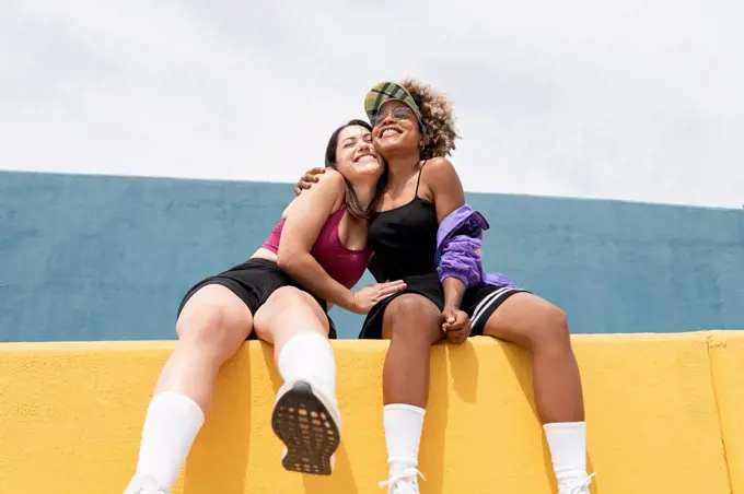 Smiling female friends embracing while sitting on yellow wall