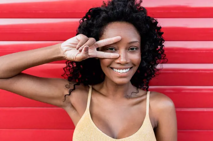 Smiling woman with curly hair gesturing peace sign in front of red shutter