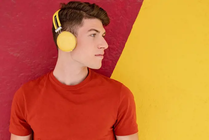 Young man listening music through wireless headphones in front of two tone wall