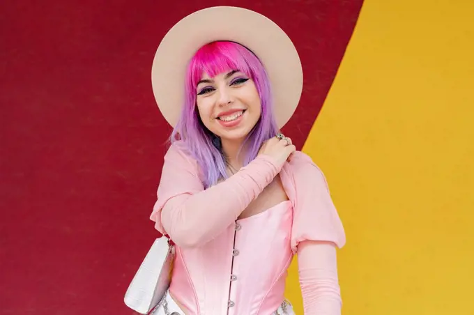 Fashionable young woman with dyed hair smiling in front of wall