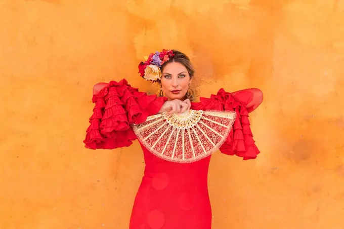 Female flamenco artist holding hand fan while standing in front of orange wall