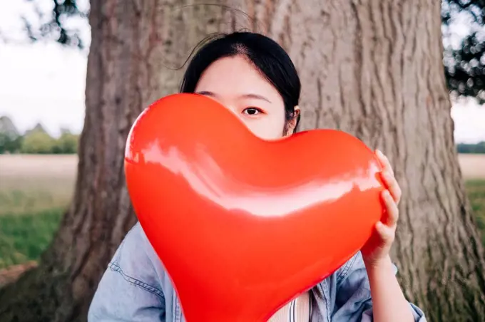 Woman covering face with red heart shaped balloon at park