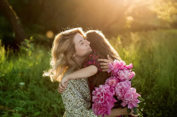 Woman with bunch of peonies embracing girl at meadow