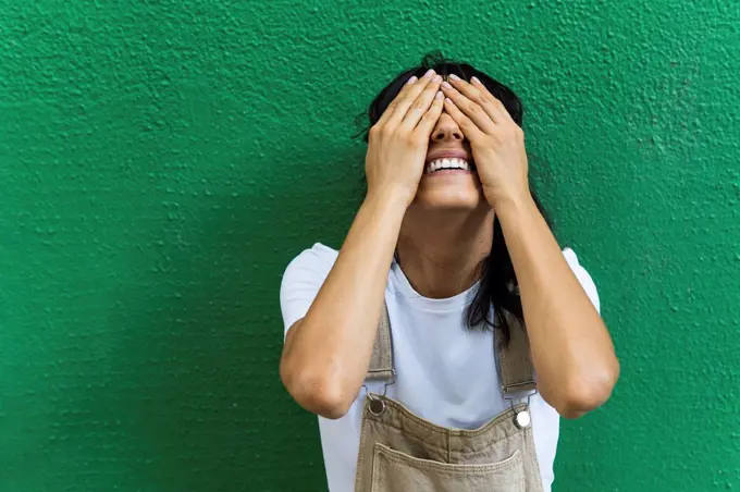 Smiling young woman covering eyes with hands in front of green wall