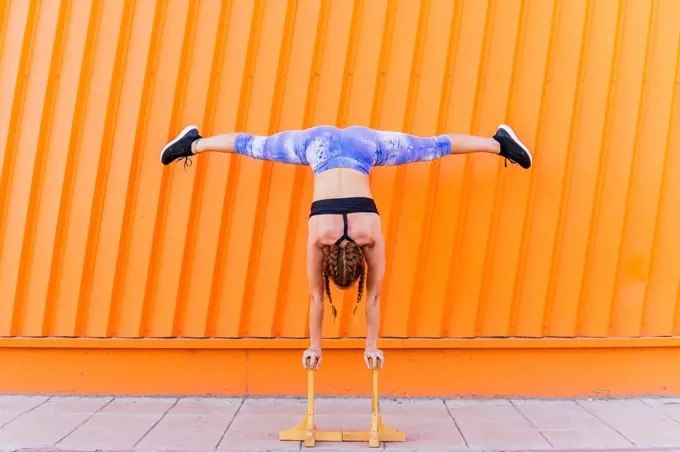 Female athlete doing handstand on sports equipment in front of orange wall