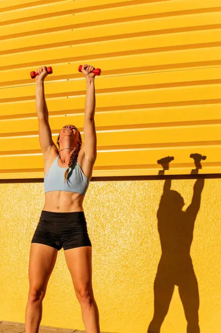 Female athlete with hands raised holding dumbbell in front of yellow wall