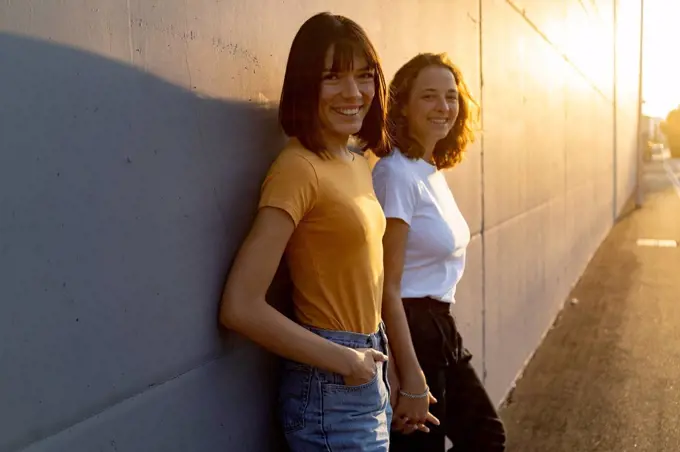 Smiling woman holding hand of girlfriend while leaning on wall