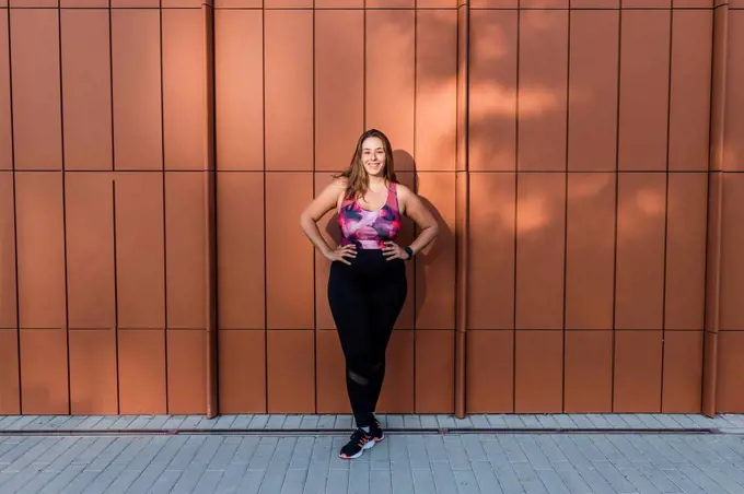 Smiling sportswoman with hand on hip standing in front of wall