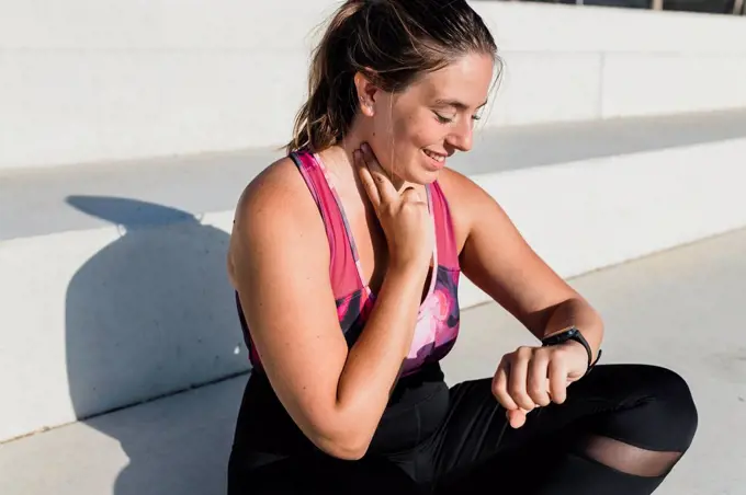 Smiling young woman looking at smart watch while checking pulse on neck during sunny day