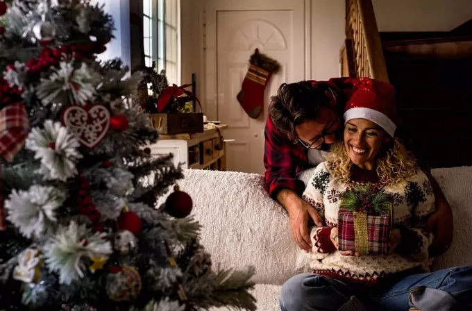 Man embracing woman holding Christmas present while sitting on sofa at home