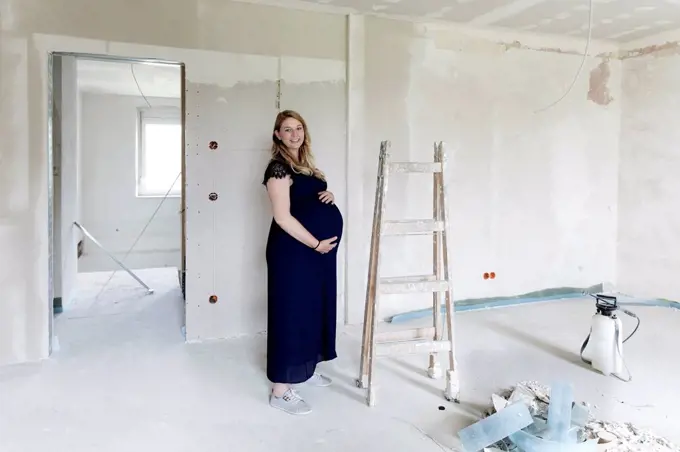 Smiling pregnant woman touching abdomen at house under renovation