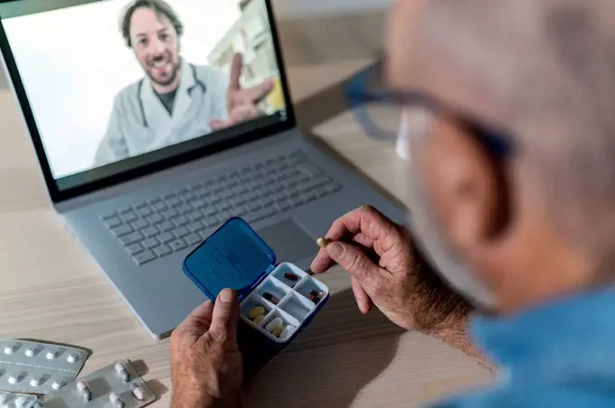 Man holding pill box on video call with doctor at home