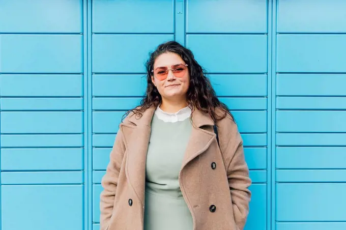 Smiling curvy woman wearing sunglasses in front of blue wall