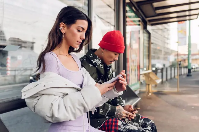 Couple using smart phones sitting on bench at bus stop