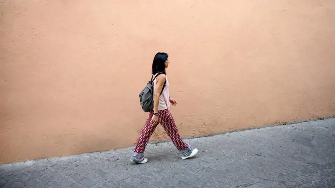 Woman with backpack waking on footpath by peach wall