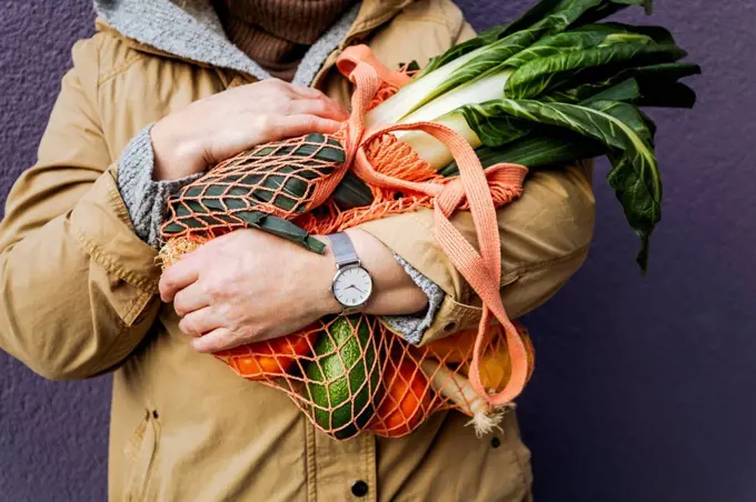 Woman holding mesh bag with vegetables in front of purple wall