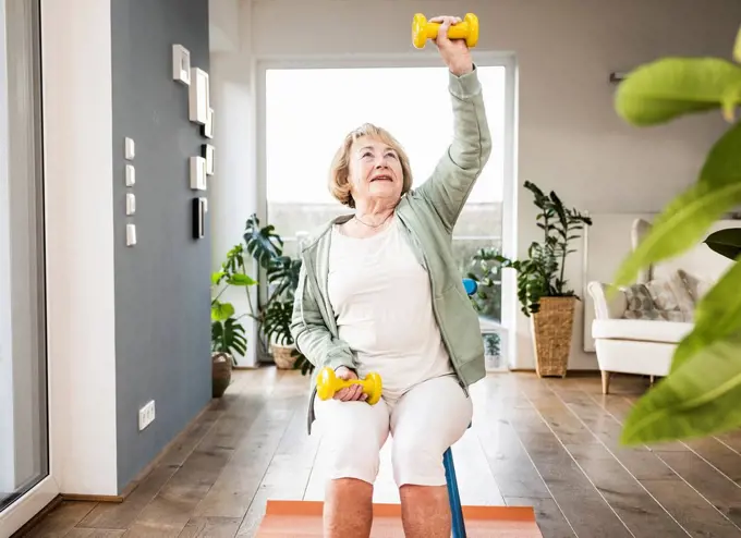 Senior woman with hand raised holding dumbbell sitting on chair at home
