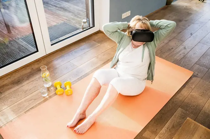Senior woman wearing virtual reality simulator exercising with hands behind head at home