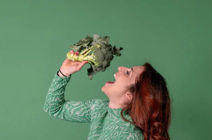 Redhead woman with mouth open holding broccoli standing against green background