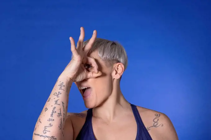 Woman with short hair looking through fingers against blue background