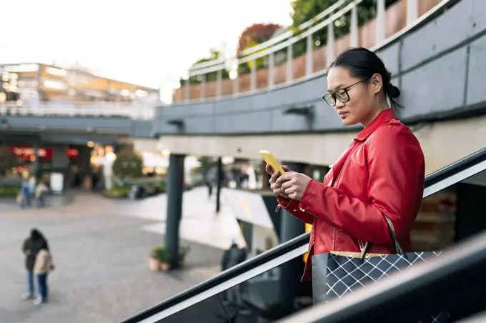 Young woman with shopping bag using smart phone moving down on escalator