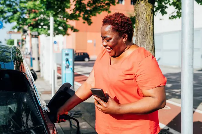 Happy woman holding mobile phone charging electric car on sunny day