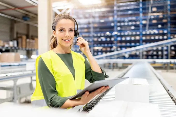 Smiling worker wearing headset standing with tablet PC by conveyor belt in warehouse