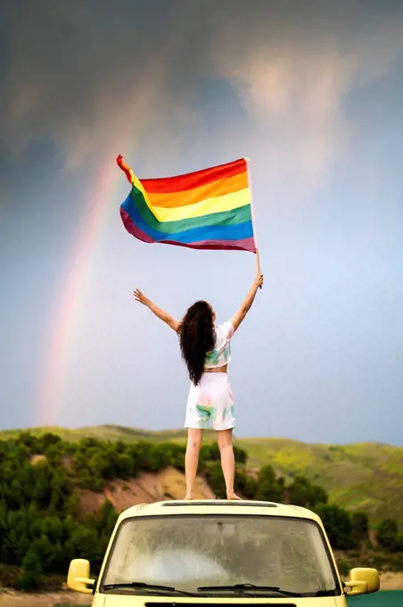 Young woman holding rainbow flag standing on van