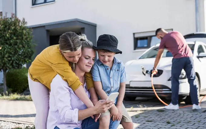 Mother and children checking smartphone while father is charging electric car