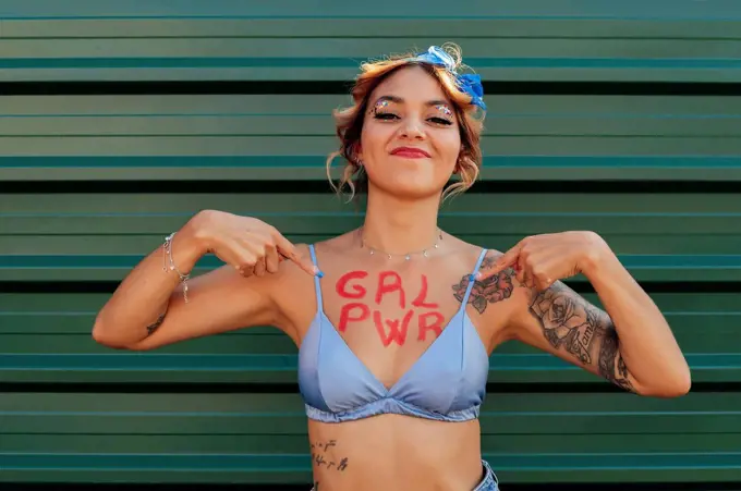 Confident woman showing girl power text on chest in front of green metal wall