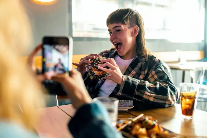 Young woman photographing girlfriend holding burger at restaurant
