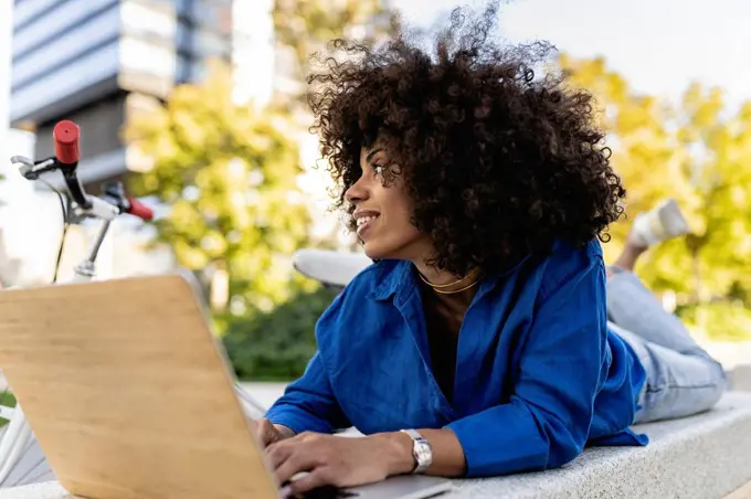 Smiling Afro woman with laptop lying on concrete bench