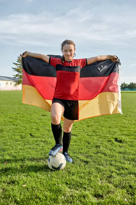 Happy girl standing with soccer ball and holding German flag on field