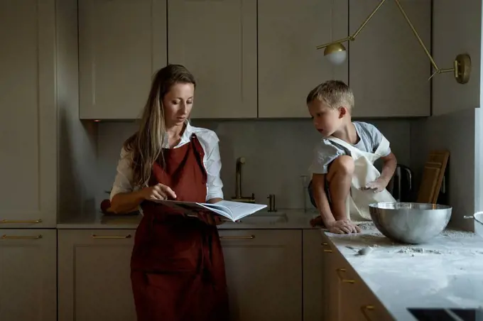 Mother reading recipe book with son sitting on kitchen counter