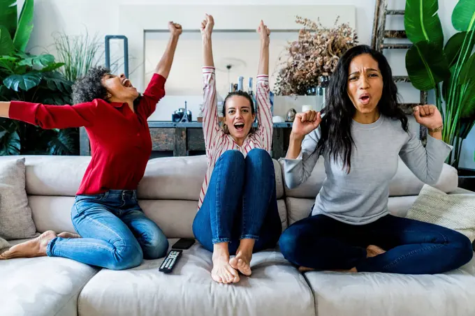 Three excited women on couch at home watching Tv and cheering