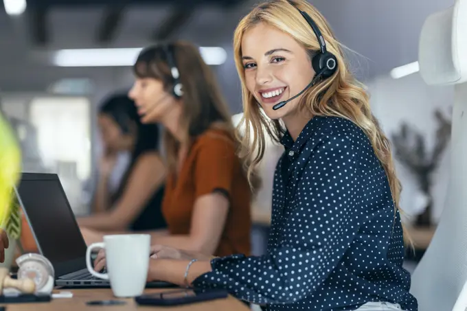 Happy blond businesswoman talking through headset sitting by colleagues at desk in office