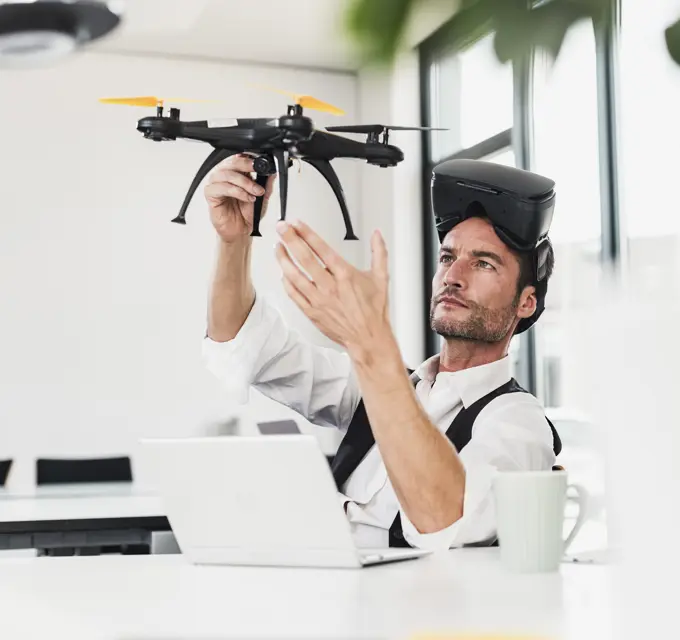 Mature businessman wearing VR glasses and examining drone at desk in office