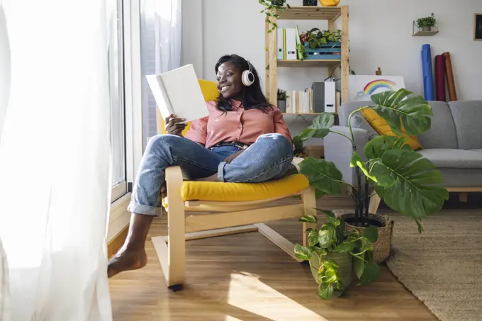 Happy woman listening to music and reading book sitting on chair at home