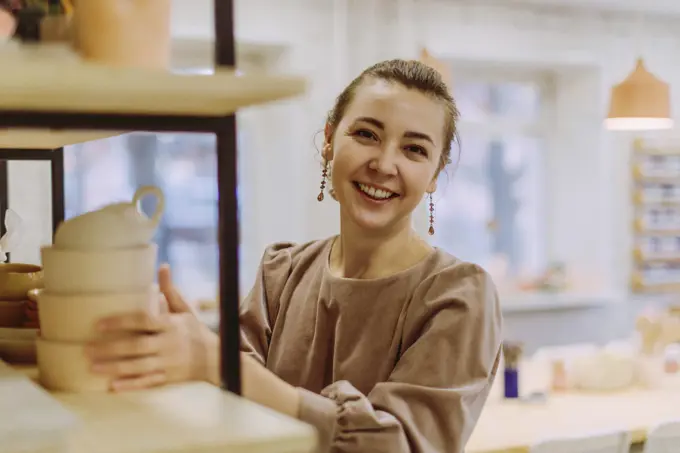 Happy businesswoman arranging handmade clay crockery in workshop