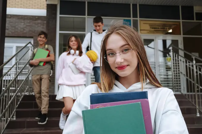 Smiling teenage girl wearing eyeglasses with books standing in front of friends at schoolyard