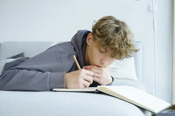 Boy lying on sofa writing in notebook at home