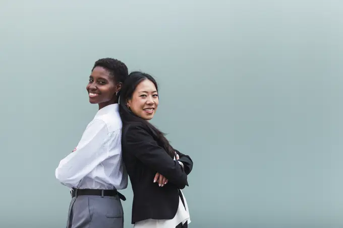 Smiling businesswomen with arms crossed standing back to back in front of blue wall