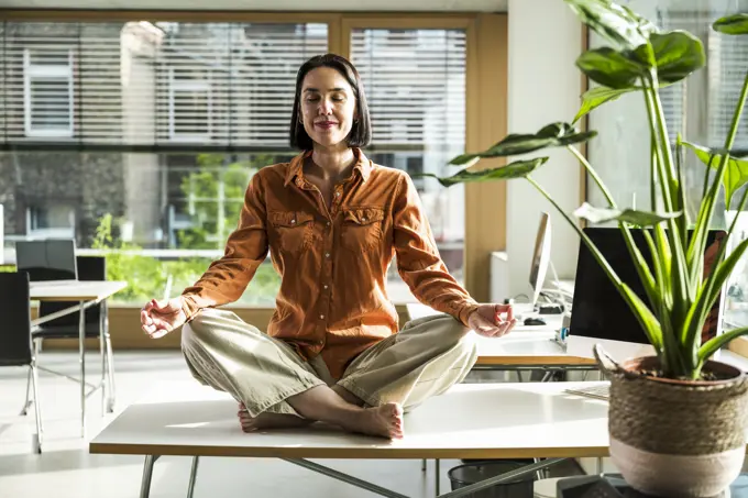 Smiling mature businesswoman meditating in lotus position on desk at office