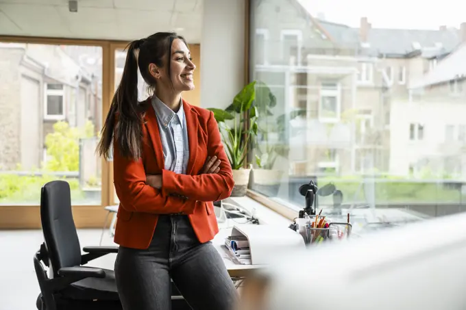 Smiling businesswoman with arms crossed looking through window in office