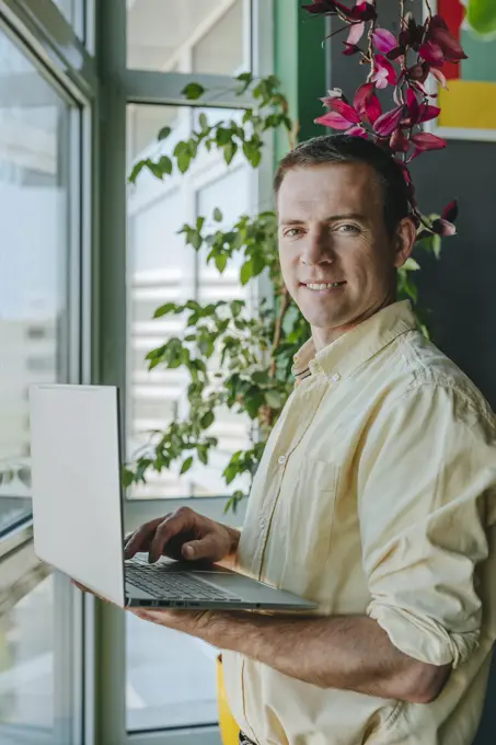 Happy businessman standing with laptop near window in office