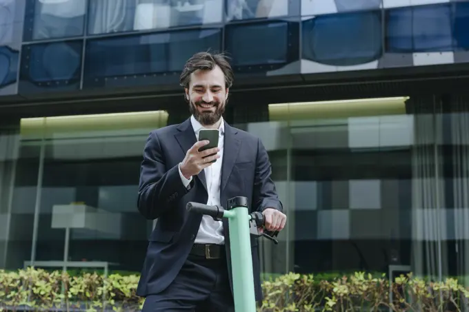 Smiling businessman standing with electric scooter and holding smart phone