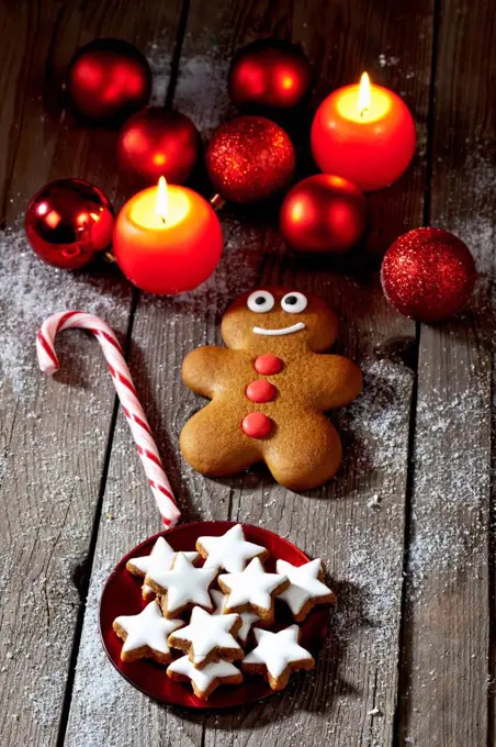 Gingerbread man, sugar cane, plate of cinnamon stars, Christmas baubles and lighted candles on grey wood