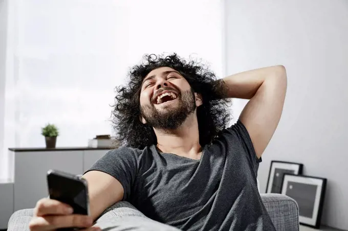 Laughing young man with smartphone sitting on couch at home