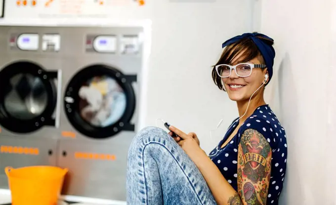 Portrait of tattooed young woman hearing music with earphones in a launderette