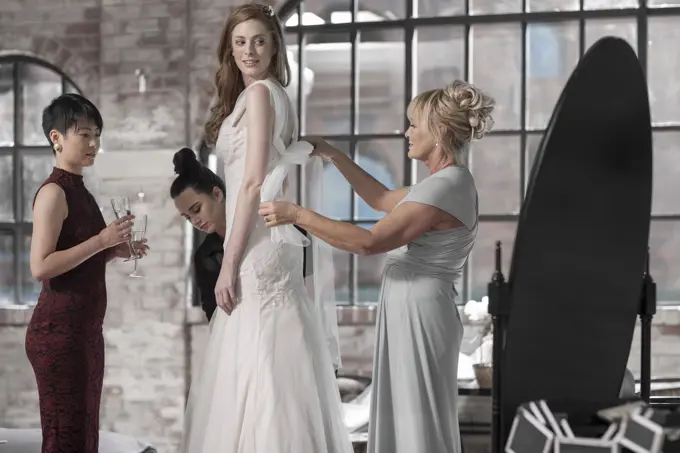 Mother and bridesmaid helping bride putting on wedding dress